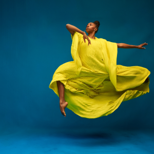 a black woman, robed in bright yellow-green, leaps and seems to float in front of a blue background