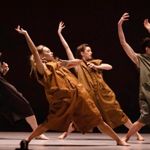 Four dancers wearing baggy rompers in earthy colors are mid-motion. Their feet are apart, and they lean back, each with an arm flung above their head. 