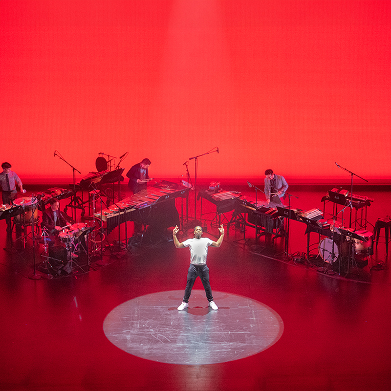 On a stage bathed in red, a single dancer stands in the center with his arms stretched upward, surrounded by mic'd percussion. 