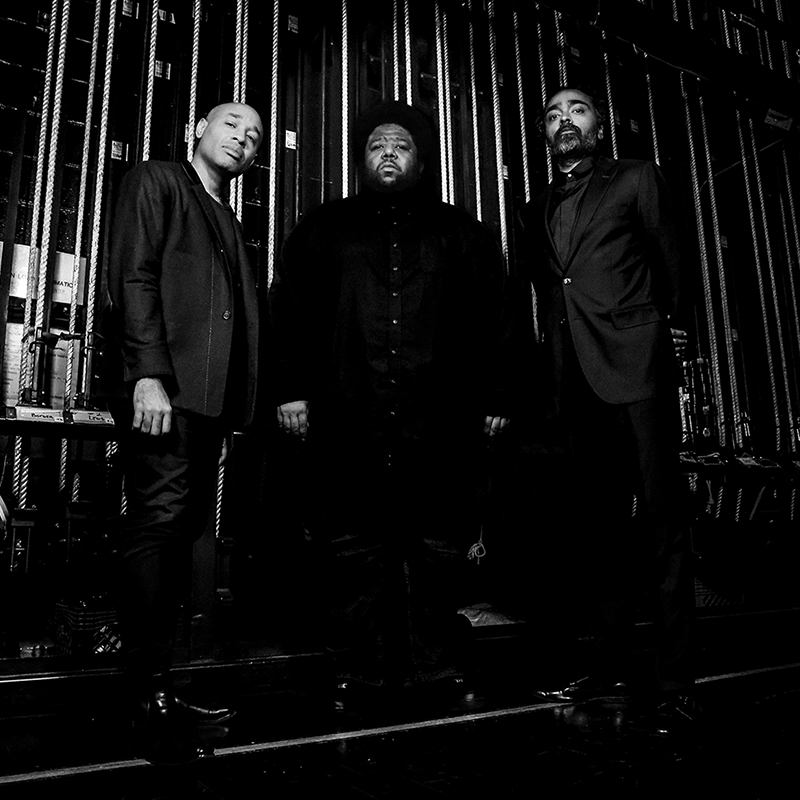 A black and white photo of three stoic black men standing in front of backstage rigging. 
