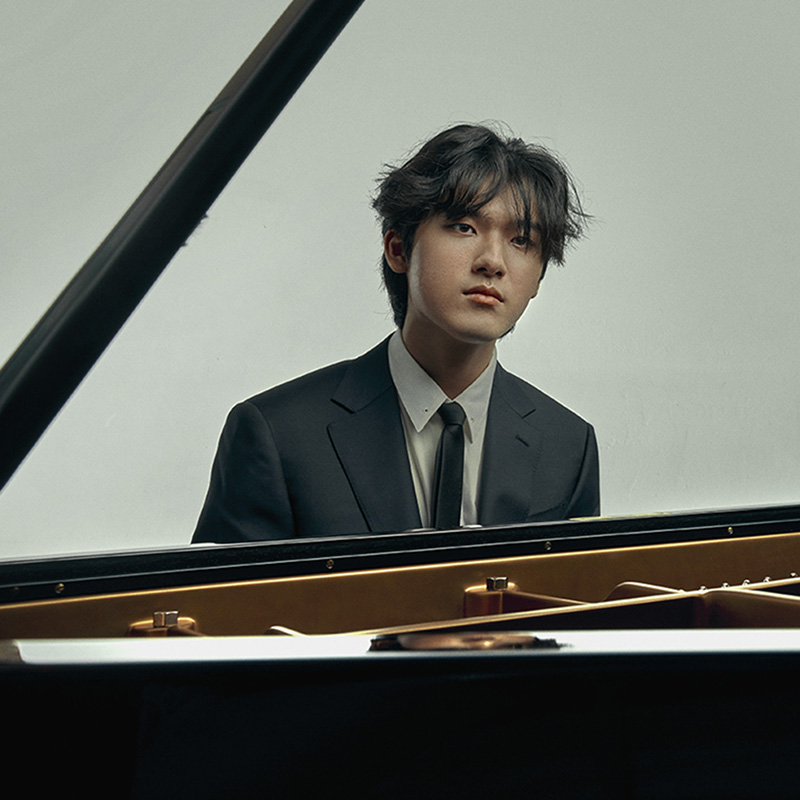 A young, South Korean man sits at the piano, his face framed by the stick of the lid, staring solemnly ahead in a dark suit. 