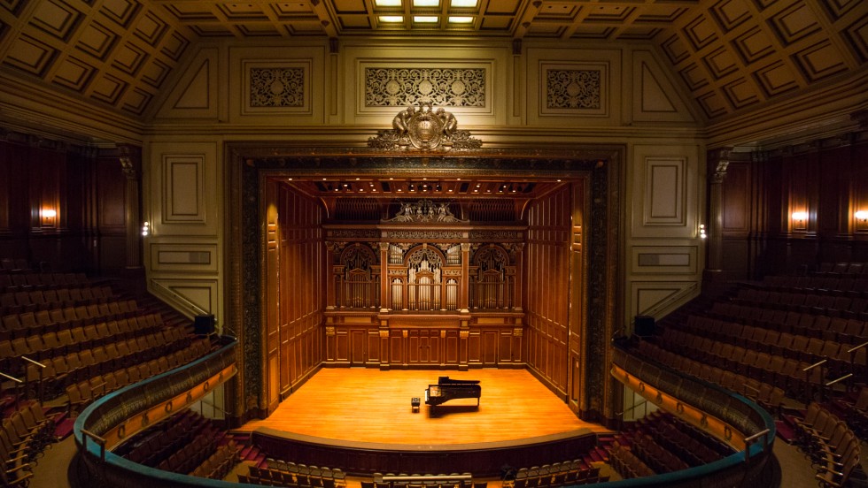 the inside of a horseshoe-shaped concert hall, with wood molding throughout and a grand piano on the stage
