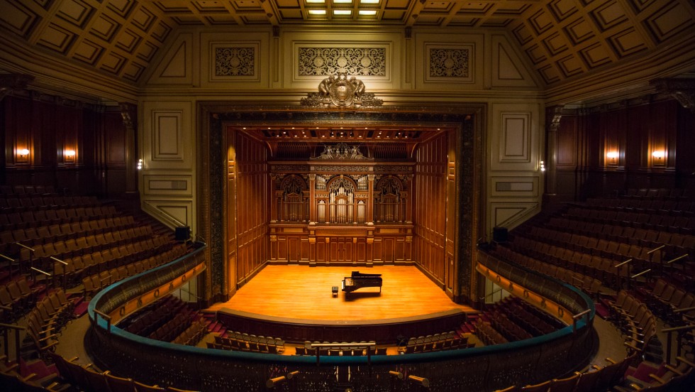 A horseshoe shaped recital hall with dark wood paneling and a ornate organ at the rear of the stage