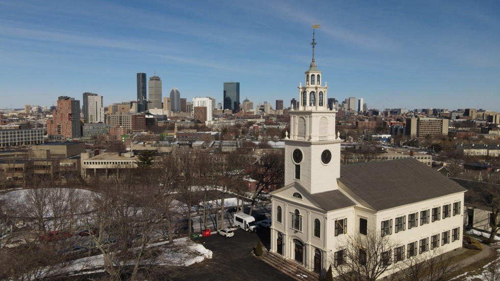 an aerial photo of a steepled, two story, white meetinghouse, with the skyline of Boston in the background