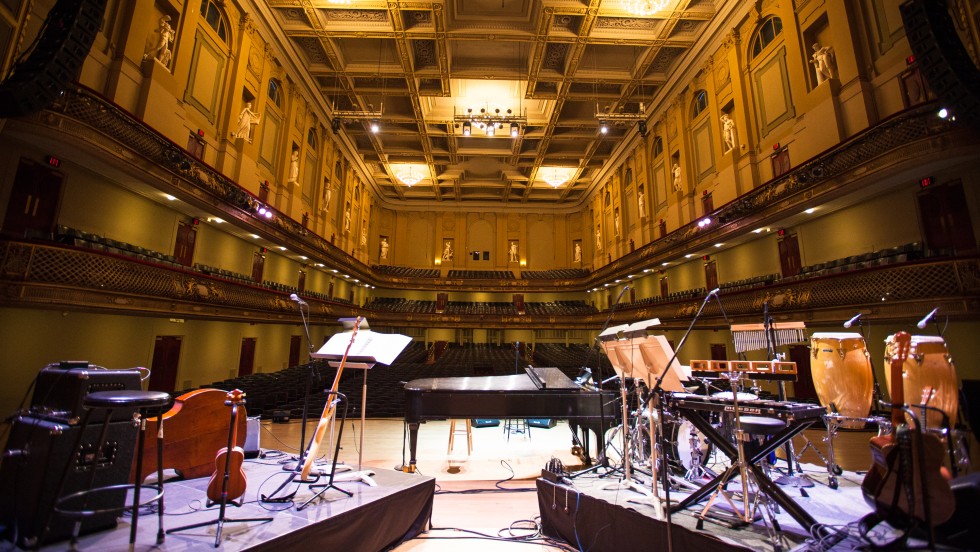 A view from the stage, an orchestra stands before a full hall of people