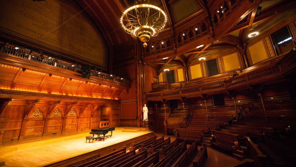 A wood paneled auditorium with a bright chandelier and golden lights illuminating dark wooden pews