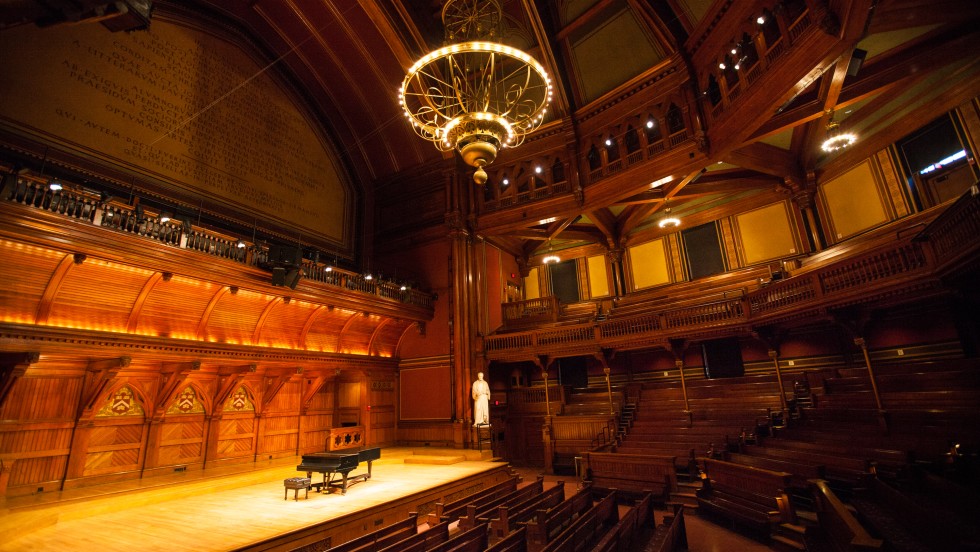 a piano sits on stage of an ornate wooden venue. a large chandelier sits above the audience.