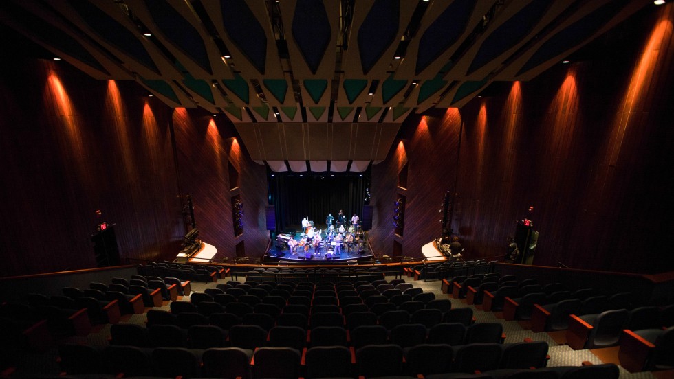 A view from the balcony, a large ensemble plays in a dimly lit auditorium