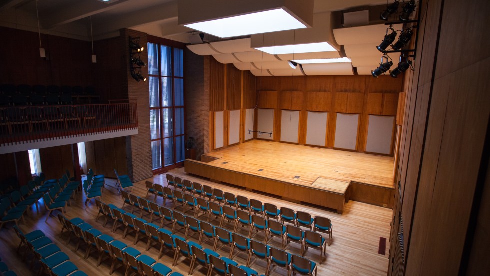 A small, wood paneled auditorium with many blue chairs