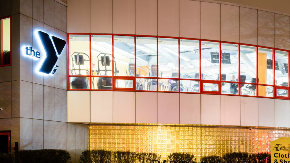 a long curved wall of glass lined with exercise machines looks out over the sidewalk. A large Y for YMCA brands the building.